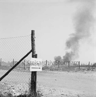 Brand: Müllkippe: vorn links Hinweisschild zur Brandstelle an Zaunpfahl: hinten rechts hinter Zaun Rauchschwaden