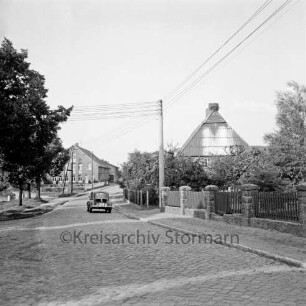 Dorfstraße mit Kopfsteinpflaster: links Bäume: dahinter Bauernhaus: in der Mitte Pkw: rechts Garten hinter Zaun: dahinter Bauernhaus mit Storchennest