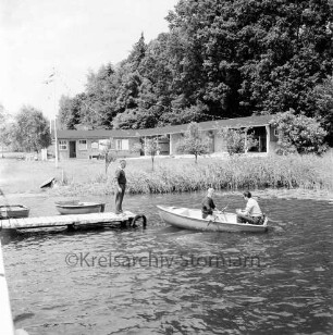 Badeanstalt: am Herrenteich: Mann auf Anleger des Badestegs: zwei Männer in Paddelboot: am Ufer Schilf, Bootshaus, Umkleideräume, Liegewiese, Fahnenmast mit Wimpelketten: im Hintergrund Waldstück