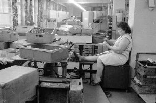 Pause im Hausschuhwerk : Wurzen. Kinderhausschuhwerk. Werkarbeiterin die zwischen den Kisten mit Kinderhausschuhen eine Pause macht, ihr Bein hochlegt und eine Apfelsine schält