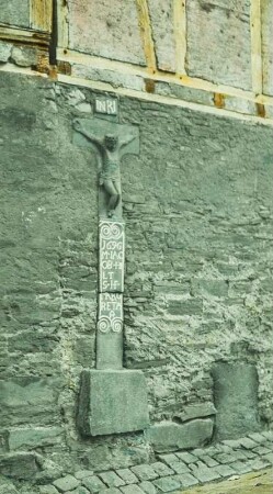 Wegkreuz : Wegkreuz, bezeichnet und datiert 1696, eingemauert in die Mauer einer Gebäudes mit Obergeschoss in Fachwerk