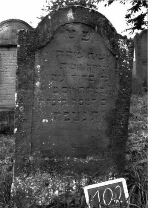 Grabstein 102 (Index: 100103529) Material: Sandstein Zustand: angewittert Maße HxBxT: 120x64x9 cm Beschreibung: Stele, eingezogener Rundbogenabschluss, Schriftfeld eingetieft mit Konvexbogenabschluss: Grabstein 102