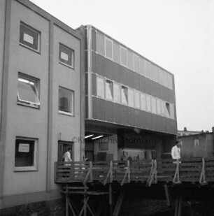 Nickel: Mühlenstraße / Lübecker Straße: Kaufhaus: Erweiterungsbau auf rückwärtigem Grundstück Mühlenstraße: Eröffnung: Außenansicht: Rückfront mit Vollverglasung, Hintereingang, Schaufenster: Fußgängerbrücke über Trave, mit Gerüst, zwei Personen: links älterer Kaufhaustrakt Nickel: Aufnahme unscharf