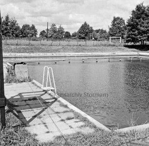 Freibad: vorn Schwimmbecken: dahinter Zaun und Sportplatz mit Fußballtor: im HIntergrund Bäume