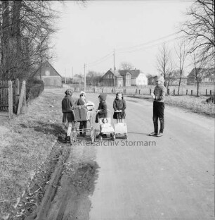 Dorfstraße: vorn Mädchengruppe mit Puppenwagen: rechts Junge: links Hecke und Büsche: hinten Bauernhäuser