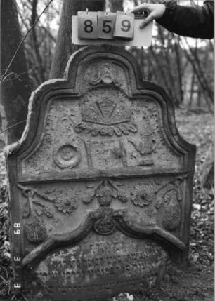 Grabstein 859 (Index: 99751239) Material: Sandstein Zustand: stark verwittert mit Textverlust Maße HxBxT: 125x87x12 cm Beschreibung: Stele, geschweifter Abschluß, Schriftfeld eingetieft Symbolik: Kanne mit Schale, aufgerollte Thorarolle, Pinienzapfen, Blumen, Muschel, Ranken: Grabstein 859