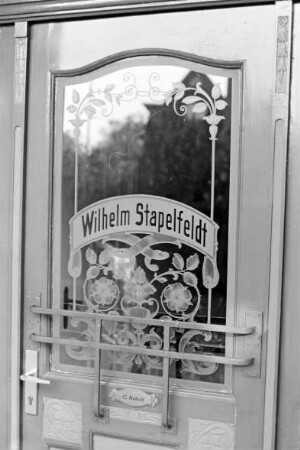 Lübecker Straße (2): Bauernhof: Wohnhaus: Eingangstür, Detailaufnahme: Glasscheibe mit Schliff und Ätzung, florale Motive, Namenszug Wilhelm Stapelfeld, davor unten Metallgitter, darunter Namensschild C. Untied