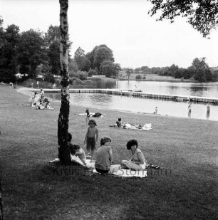 Großensee: Südufer: Freibad: vorn Liegewiese: darauf Gruppen von Besuchern: vorn links Baum: hinten Holzstege im Wasser: davor Nichtschwimmerbereich: hinten links Campingplatz und Bäume