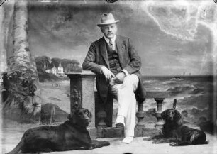 Porträt eines Mannes mit zwei Hunden vor gemaltem Hintergrund : Mann mit zwei Hunden auf einer Balustrade sitzend vor gemaltem Hintergrund, die Ostsee mit Alexandrinen-Cottage an der Küste in Heiligendamm zeigend, Atelier Beckmann in Heiligendamm.