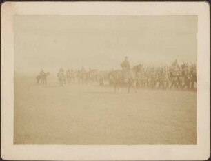 Berittene Uniformierte und vorbeiziehende Infanterie.