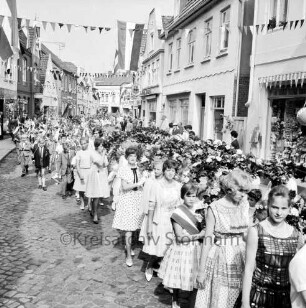 Kindervogelschießen: Umzug: Hindenburgstraße: ältere Mädchen mit Blumenbögen, dahinter Jungen mit Blumenstäben und Fähnchen: an den Straßenrändern Zuschauer: über Straße Wimpelgirlanden, an Fassaden Fahnenschmuck: an linker Straßenseite Friseursalon Lübcke, hinten Gaststätte "Bierquelle": an rechter Straßenseite hinten Schuhgeschäft Paul Jürgens: im Hintergrund Hagenstraße, Gaststätte "Stadt-Krug", Toreinfahrt Holz- und Baustoffhandlung Hugo Eggers, 2. Juli 1963