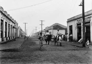 Straßenszene in Nova Granada : Häuser mit Geschäften, Bürgersteige und Strommasten rechts und links einer Straße in Nova Granada. Auf der Straße Reiter und Passanten.