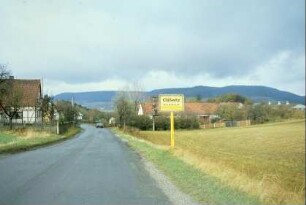 Straße mit Ortsschild, rechts Ackerfläche; im Hintergrund