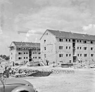 Klosterbergen: Bogenstraße: Wohnblocks im Rohbau, links Dachdeckerarbeiten: vorn links Front eines VW-Käfers: dahinter Bretterhaufen, Rohre sowie Baumaterial: dazwischen Arbeiter: hinten links Einfamilienhaus