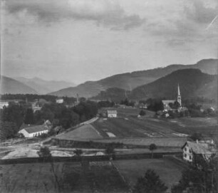 Dorf : Blick auf ein Dorf in einer Hochebene. Im Hintergrund bewaldete Berghänge und eine Kirche, im Vordergrund Felder und Weiden