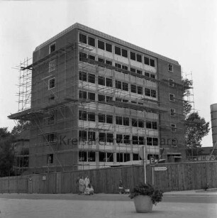 Hochhaus [später Gebäude B]: Mommsenstraße: Rohbau mit Gerüst: davor Bauzaun, wei Frauen mit Kinderwagen und Kind mit Puppenwagen: rechts Betonsilo: dahinter Hauptgebäude [später Gebäude A]: vorn rechts Blumenschale