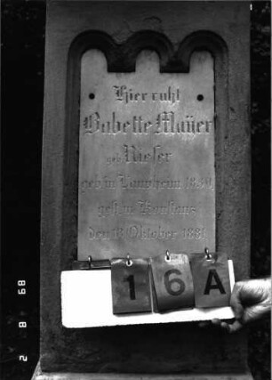 Grabstein 16 (Index: 100101734) Material: Sandstein, weiße Marmorplatte Zustand: unten abgeblättert, Aufsatz fehlt Maße HxBxT: 154x50x38 cm Beschreibung: konische Stele, profilierter Sockel, Abdeckplatte mit Aufsatz, Schriftplatte mit dreifachem Rundbogenfries Symbolik: gestelztes Rundbogenfries Steinmetz: Epple: Grabstein 16A