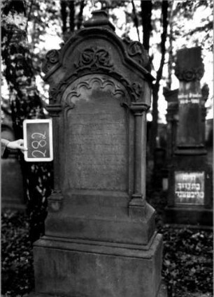 Grabstein 282 (Index: 100112050) Material: Sandstein Zustand: verwittert Maße HxBxT: 190x64x30 cm Beschreibung: ädikulaartige Stele mit Säulen, getreppter Sockel, schräg eingezogener Segmentbogenabschluß mit Mittel- und Eckakroteren, Schriftfeld eingetieft mit Rundbogenabschluß Symbolik: Ranken, Rundbogenfries: Grabstein 282