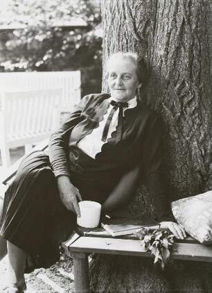 Ältere Dame mit Kaffeetasse und dem Buch "Frühling im Dorf" von Hans Sterneder auf der Rundbank um einen Baum in einem parkähnlichen Garten sitzend