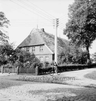 Dorfstraße mit Kopfsteinpflaster und Fahrradfahrerin: dahinter Strommast: rechts Baum: hinten Mitte Bauerngehöft Karl Thormälen mit Vorgarten und Hecke