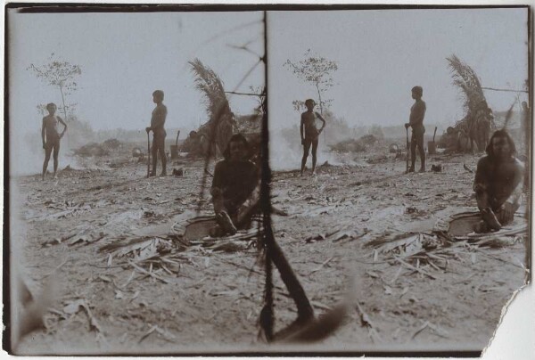 Kitchen area of a Kayapó village, in the foreground a man working with feathers