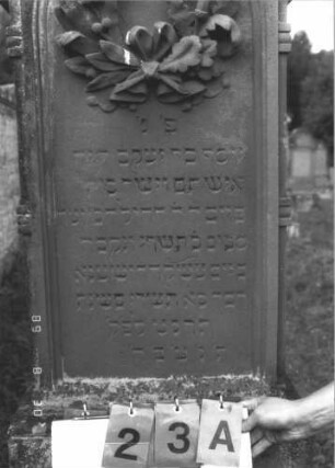 Grabstein 23 (Index: 99609659) Material: Sandstein Zustand: verwittert Maße HxBxT: 189x63x24 cm Beschreibung: Stele, Segmentbogenabschluß, in Voluten auslaufend, Volutenaufsatz, Blüte, Schriftfeld eingetieft, getreppter Sockel Symbolik: Kranz, Blüte: Grabstein 23A