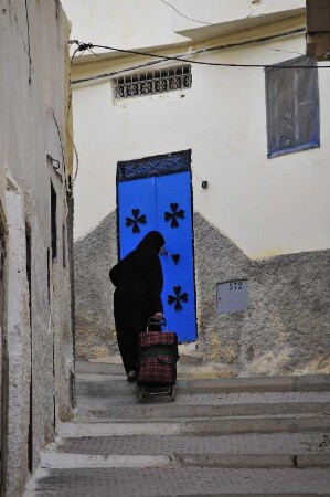 Moulay Idriss - Schwarz verhüllte Frau vor einer blauen Haustür : Eine schwarz verhüllte Frau transportiert eine Last mit einem kleinen Kofferwagen die Treppen bergan. Aufgenommen am 30. Oktober 2010 in Moulay Idriss