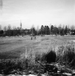 Groten Diek: Eisdecke: in der Mitte zwei Kinder, eines mit Fahrrad auf Teich: vorn Schilf: am gegenüberliegenden Ufer Turm der Auferstehungskirche, Häuser und Bäume: Winter