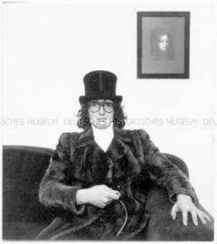Junger Mann mit Pelzmantel, Brille und Zylinder auf dem Sofa sitzend, fotografiert sich mit Selbstauslöser