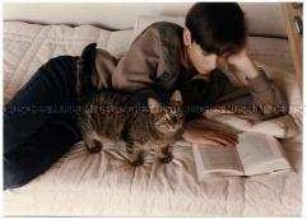 Junge liest auf dem Bett, Katze steht daneben (Altersgruppe bis 14)