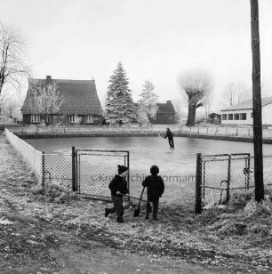 Dorfteich: im Vordergrund zwei Kinder an Zaundurchlass mit Eishockeyschlägern: Schlittschuhläufer auf Teich: im Hintergrund Bäume und Häuser: Winter