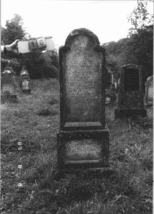Grabstein 16 (Index: 99799585) Material: Sandstein Zustand: stark verwittert Maße HxBxT: 170x55x28 cm Beschreibung: Stele, schräg eingezogener Rundbogenabschluß, Schriftfeld eingetieft, getreppter Sockel mit eingetieftem Schriftfeld Symbolik: floral: Grabstein 16