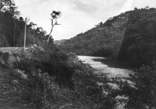 Flusslandschaft in Brasilien : Ein Fluss verläuft zwischen einem bewaldeten Hügel rechts und einer Straße auf der linken Seite.