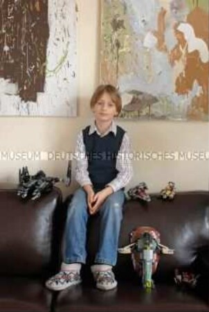 Junge auf dem Sofa sitzend mit Fantasy-Legofiguren und abstrakten Gemälden im Hintergrund (Altersgruppe B)