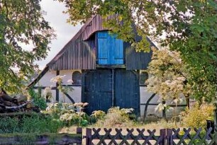 Glashütte: Fachwerkkate mit Wellblechdach: Giebelseite Wirtschaftstrakt mit Scheunentor, darüber Heuluke: links Holzstapel, vorne Holzzaun, Pforte