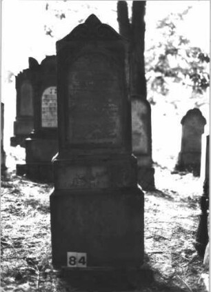 Grabstein 84 (Index: 99898849) Material: Sandstein Zustand: stark verwittert Maße HxBxT: 142x52x24 cm Beschreibung: aufgesockelte Stele, Konvexbogenabschluss mit Voluten und Mittelpalmette Symbolik: Voluten, Palmette: Grabstein 84