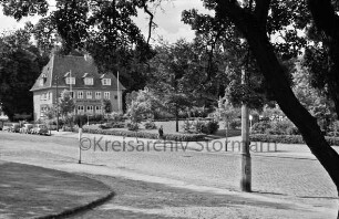 Sülzberg: Kurpark: vorn Fußgängerweg mit Straßenlampe und Baum: hinten Mitte Rondeel mit Bänken, Rabatten und Hecken: hinten links Haus des Handwerks: davor parkende Pkws: im Hintergrund Bäume