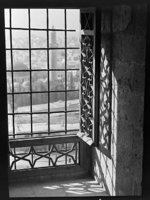Blick durch ein vergittertes Fenster auf eine Stadt, im Mittelgrund eine Moschee