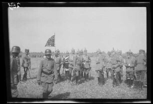 Bei Focșani (Rumänien): Gruppe von Offizieren mit Pickelhaube neben einer Standarte mit Tatzenkreuz auf einem weiten Feld, im Vordergrund ein abmarschierender Soldat mit Stahlhelm, im Hintergrund große Ansammlung von Soldaten ("Kaiserparade")