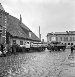Verkehrsunfall: Lübecker Straße: Lkw fährt in Haus: Unfall-Lkw der Meierei-Genossenschaft Reinfeld mit Anhänger, darauf Milchkannen, mit Kühlerschnauze in Fassade von Eckhaus zu Lübecker Torbrücke: Person läuft über Straße: links auf Bürgersteig Schaulustige, an Straßenrand Fahrradanhänger mit Milchkannen: rechts auf Bürgersteig Junge, Schaulustige: Straße mit nassem Kopfsteinpflaster, Lichtmast: hinten Straße Pferdemarkt, Eckhaus zur Lübecker Torbrücke, daran Firmenname F. Bartheidel, Werbeschrift "Gasolin": im Hintergrund Gewerbebetrieb, Schornstein