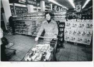 Frau mit traurigem Gesichtsausdruck mit einem gefüllten Einkaufswagen im Supermarkt (Altersgruppe 18-21)