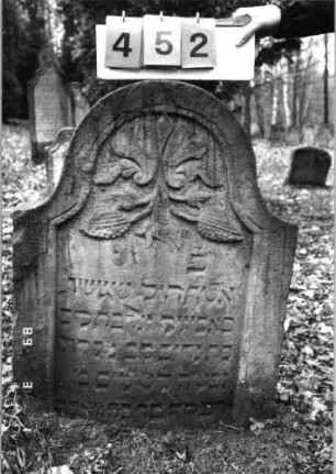 Grabstein 452 (Index: 99752824) Material: Sandstein Zustand: gut, Stele eingesunken Maße HxBxT: 88x66x9 cm Beschreibung: Stele, eingezogener Rundbogenabschluß, Schriftfeld eingetieft Symbolik: Pinienzapfen, Lilie Besonderheit: Zitat Proverbia 12, 4 und 31, 10: Grabstein 452