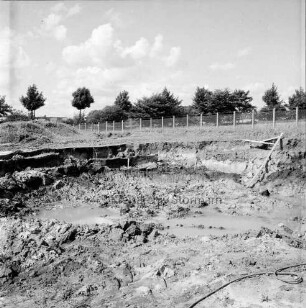Baugrube mit Wasserlöchern: hinten Zaun und Baumreihe