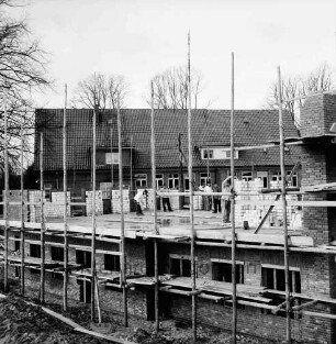 Volksschule: Erweiterungsbau für 3. Klasse: Grundmauern mit Gerüsten: darauf Arbeiter: dahinter altes Schulgebäude und Bäume