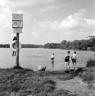 Großensee: Südufer: Badestelle: vorn links Hinweisschilder und Rettungsring: in der Mitte Findling: rechts am Ufer und im Wasser Frau, Junge und Mann: hinten Mitte weitere Schwimmer: rechts am gegenüberliegenden Ufer hinter Bäumen Zelte