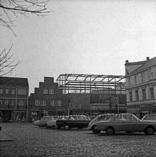 Firma Carl Hoffmann: Hindenburgstraße: Elektrohandel: Neubau: Aufbau des Stahlgerüsts: davor Bauzaun: links Wohn- und Geschäftshauser des Haushaltswarengeschäfts Otto Möller und Lamprecht: rechts Hausecke des Wohn- und Geschäftshauses mit Drogerie Purnhagen: im Vordergrund Marktplatz und parkende Pkws