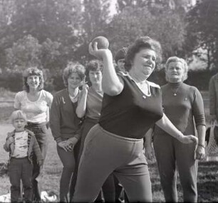 Kugelstoßen : Sportfest des Deutschen Turn- und Sportbundes (DTSB) in der DDR, zur Leibesertüchtigung der Bevölkerung. Kräftige Frau, um die 60 Jahre, bemüht sich durch das Kugelstoßen ein weiteres DDR-Sportabzeichen zu erlangen. Im Hintergrund stehen weitere Frauen, von Jung bis Alt, die sich über das Geschehen im Vordergund amüsieren beziehungsweise mitfiebern