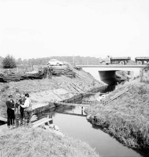 Beste: Flussregulierung: Zusammenfluss von links Norderbeste und vorn rechts Süderbeste bei Blumendorf: Böschungserneuerung mit Bohlenwänden und Ziegelbrocken: vorn links Behelfsteg über Norderbeste: darauf drei Arbeiter beim Rammen der Holzpfähle: dahinter Stapel mit Holzpfählen auf Uferböschung: dazwischen zwei Arbeiter: hinten Mitte weitere Steg über Beste: hinten rechts Brücke der Bundesstraße B 404 mit Pkws und Lkw