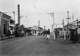 Straßenszene : Eine belebte Straße in Salto de Ytú. In der Bildmitte sind zahlreiche Männer versammelt. Rechts am Bildrand auch einige Frauen. An der Straße stehen Masten mit Laternen und Leitungen.