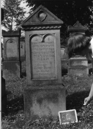 Grabstein 54 (Index: 99801834) Material: Sandstein Zustand: Giebel verwittert Maße HxBxT: 170x50x21 cm Beschreibung: Stele, Sockel, Giebel, eingetieftes Schriftfeld mit doppeltem Rundbogen Symbolik: verwitterte oder abgefallene Blume ? im Giebel, Zahnfries: Grabstein 54